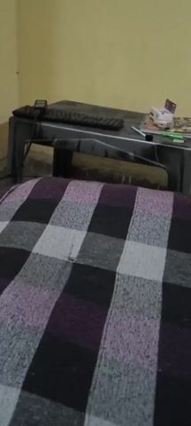 The video shows a dimly lit room from a low, stationary perspective, likely from a bed covered with a purple, gray, and black checkered blanket. In the background, a black metal desk is visible, with a keyboard and a small device on it. There are also some items on the desk, including what appears to be a small white box and some books or papers. To the right of the desk, there's a wall with a yellow-toned paint. Further in the background, a doorway leads to another area where two large blue water jugs are standing. The overall environment appears to be a residential or dorm room.
