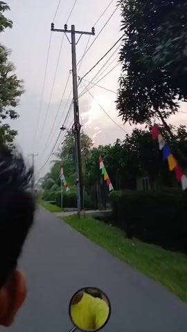 The video shows a first-person perspective of someone riding a motorcycle down a paved road. The sky is overcast with the sun peeking through the clouds. There are power lines running overhead, supported by utility poles. On the right side of the road, there is lush green vegetation, including trees and bushes. There are also several small flags with red and white colors, which appear to be Indonesian flags, attached to poles along the roadside. On the left side of the road, there are houses and more vegetation. In the distance, another motorcycle can be seen traveling in the same direction. The rider's left hand is visible in the lower left corner of the frame, and a portion of the motorcycle's rearview mirror is visible in the lower center of the frame, reflecting a yellow object. The overall scene suggests a rural or suburban setting during daylight hours, possibly late afternoon or early morning due to the lighting conditions.