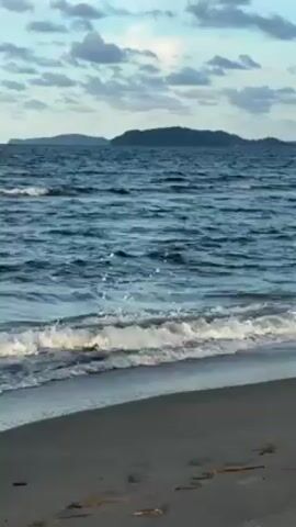 The video shows a beach in Tela, Honduras. The beach has dark sand and a calm ocean with small waves gently lapping at the shore. In the distance, there are two islands. The sky is partly cloudy. The video appears to have been taken at dusk or dawn.