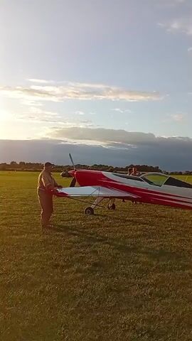 A person in a camouflage jacket and brown pants is standing on the grass beside a red and white small airplane with its propeller spinning. Two other people are visible near the cockpit of the airplane. The sky is blue with some clouds, and the sun is low on the horizon, casting a warm light. The airplane is on a grassy field.