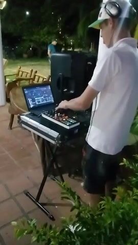 Here is a description of the video:

A young man wearing a white t-shirt, dark shorts, a white baseball cap with a green brim, and headphones is standing behind a DJ setup on a red brick patio at night. He is operating the mixing console. A laptop displaying a DJ interface is next to the console. A speaker is positioned directly behind the DJ setup. Behind him is a partially visible wooden structure supported by white columns, a few chairs, and some greenery. In the background, another person is standing. The overall lighting is dim.