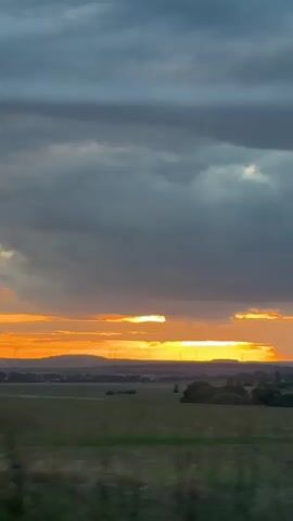 The video shows a view from a vehicle moving along a rural road at dusk. The sky is filled with dramatic, dark clouds with a band of golden-orange sunset visible near the horizon. The landscape consists of open fields, some with crops, and scattered trees. In the distance, there are signs of human habitation with buildings and structures forming a village or town. There are also some wind turbines visible on the horizon. Road signs are present along the side of the road, including what appear to be prohibition signs. The road itself is paved and has white lane markings. The overall impression is a serene, yet somewhat moody, rural scene at the end of the day.