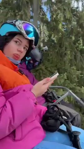 Here is a description of the video:
The video shows two individuals riding a ski lift. The camera is likely handheld, providing a first-person view from the chairlift. One person, presumably the videographer, is in the foreground wearing a pink and orange jacket, blue pants, and a blue helmet with goggles. They are looking at a smartphone. Another person is sitting next to them, wearing a purple jacket and a dark helmet. The chairlift passes over a snowy ski slope with scattered patches of green vegetation. Tall evergreen trees line the edges of the ski run. The weather appears sunny, with the sun visible through the trees. The video shows other ski lifts and skiers on the slope.