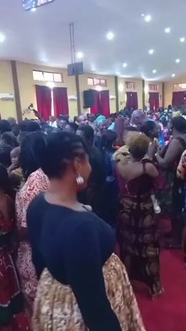 The video shows a large, crowded indoor gathering in Ogoja, Nigeria. Numerous individuals, including adults and children, fill a spacious hall with light brown walls and red curtains adorning windows or archways. Overhead lighting fixtures illuminate the room, and large speakers are mounted high on the walls, suggesting an event with amplified sound. Air conditioning units are also visible on the walls.

People are standing close together, many wearing vibrant clothing, including traditional African attire. Some individuals, like a woman in a maroon top holding a baby, and others in patterned dresses and skirts, are prominent in the foreground. A man in a blue shirt is seen standing on an elevated platform or area in the background, observing the crowd and smiling, suggesting he might be a speaker, host, or leader of the event.

The atmosphere appears lively and communal, with people mingling and looking engaged. The specific nature of the event is not explicitly stated, but it resembles a social gathering, possibly a church service, concert, or celebration. People appear to be in good spirits, and the overall scene conveys a sense of positive energy and community engagement. No visible text is present in the video.