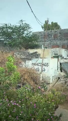 The video shows a panning view of a street in a village. In the foreground, there are several objects scattered on the ground, including stones, branches, and what appears to be debris. There are also a few plants and flowers visible. Further down the street, there are houses with various structures and materials. People are seen sitting outside their homes and interacting with each other. Some motorcycles are parked along the street. In the background, there are trees and a hill. The overall impression is that of a humble and somewhat neglected rural environment.