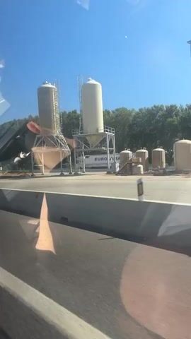 The video shows a view from a vehicle driving along a highway. On the right side of the road, there are two large industrial silos, one taller than the other, mounted on metal frames. A truck with the word "EUROLOMA" written on its side is parked in front of the silos. Further down the road, there is a small white structure with a window and a pitched roof. A light pole stands beside the road, and beyond that, a dense line of trees forms a backdrop against a clear blue sky. The vehicle is moving, causing the scenery to blur slightly. Reflections are visible on the window.