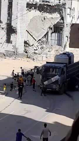 A group of people, including children and adults, are gathered around a large truck. The truck has several large water tanks in its bed and is parked on a street in front of a partially destroyed building. Some people are carrying buckets and containers, and some appear to be interacting with the truck or its driver. The building in the background is heavily damaged, with sections of walls and debris visible. The ground appears to be paved. The overall scene suggests an effort to distribute or collect water amidst a damaged environment.