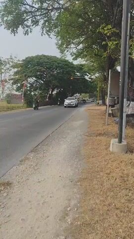 A white SUV is driving down a paved road lined with dry grass and trees. A person in a red, white, and purple shirt and green pants walks along the side of the road carrying two bags. Another person on a motorcycle drives by in the background. The sky is overcast.