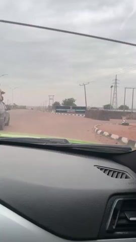 The video shows a view from the dashboard of a car driving towards the entrance of the Nigerian Defence Academy in Kaduna, Nigeria. The sign indicating the Academy's entrance is visible, framed by a cloudy sky and utility poles. The car then turns right onto a dirt road. There are no people visible within the video's frame. The audio features music in the background.