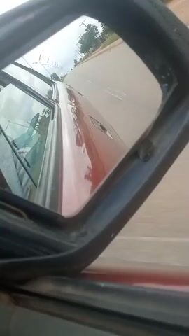 The video captures a rear-view mirror's reflection of a red car driving on a paved road. The road is lined with trees and appears to be in a rural or suburban area. The sky is overcast. The interior of the car is partially visible through the side window, including the driver's side windshield wipers. The rear-view mirror itself is framed by black rubber and is attached to the car's exterior. The car is moving, indicated by the blurred background. There are no people clearly visible in the reflection. The video is shot from inside the car, looking out towards the rear-view mirror.