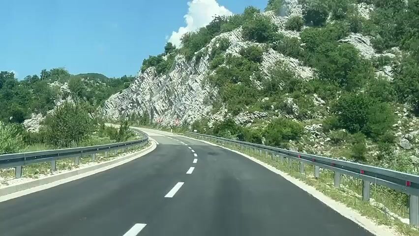 The media file captures a first-person perspective of a vehicle driving on a winding road through a mountainous, green landscape. The road is paved and has white lane markings, with a metal guardrail on both sides. The terrain is characterized by steep, rocky hillsides covered in dense green vegetation and trees. The sky is clear and blue, suggesting a sunny day with fair weather. There are a few traffic signs visible on the side of the road, including warning signs with triangular symbols. The time of day appears to be daytime, likely late morning or early afternoon, given the bright sunlight and shadows. The scene evokes a sense of travel and exploration through a natural, scenic environment. No people are visible in the frame.