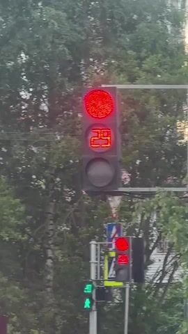 The video shows a traffic scene in Murino, Russia, with a red traffic light displaying a countdown timer. The camera is positioned behind a red Ford Ecosport. In front of the Ford, a person on a scooter with an "Ozon fresh" delivery bag is waiting at the red light. The traffic light above the scooter also shows a red light and a countdown timer. Other traffic signs are visible, including a "STOP" sign and a circular sign indicating a 24-ton weight limit with an arrow pointing to the right. The scene appears to be taking place during the daytime, with overcast skies, suggesting it could be a cloudy day or early evening. Trees line the background, and a building is partially visible to the right. The overall mood is that of a typical urban traffic situation. The license plate of the red car is visible, reading "B300HX 198". The scooter's license plate is also visible, reading "С П 5197".