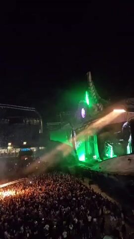 The video shows a large outdoor concert at night. A massive stage set designed to resemble a dragon is illuminated with green and purple lights. A plume of fire erupts from the stage, aimed towards a huge crowd of people. The crowd, a dense mass of individuals, is cheering and holding up their phones, their lights creating a sparkling effect. The overall atmosphere is energetic and visually spectacular, with dramatic lighting and pyrotechnics.