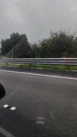 The video appears to show a roadside scene in San Miguel Topilejo, Mexico, under overcast and possibly rainy weather conditions. The perspective is from inside a moving vehicle, looking out.

In the first few seconds, an orange traffic cone is visible on the shoulder of the road. Shortly after, a white SUV is parked on the side of the road, with two individuals standing near it. A white police vehicle with the text "GUARDIA NACIONAL" and a checkered pattern along its side, featuring an emblem, drives by. A yellow diamond-shaped road sign with a black arrow indicating a left turn is also visible. Further along the road, a brick wall with graffiti runs parallel to the highway. A billboard with a picture of a man and text in Spanish, including "CHUCHOS POR EL CAMBIO" and "VERDE," is seen in the distance. The overall scene suggests a routine road passage, possibly with some minor traffic incident or police presence. The sky is uniformly gray, indicating a cloudy or overcast day, and the road surface appears wet, further suggesting recent or ongoing rain.