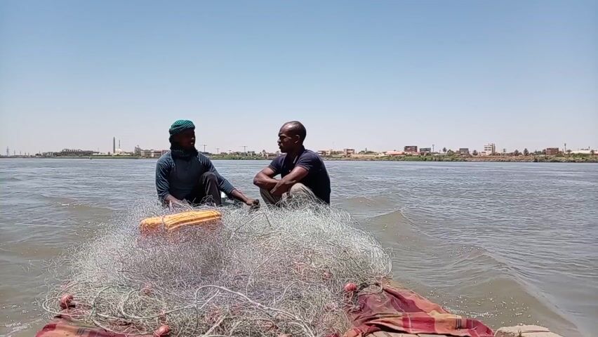 Two men are on a small, red and brown wooden boat on a wide, light-brown river. They are sitting near a large pile of gray fishing net with orange floats attached. The man on the left wears a dark-blue long-sleeved shirt and a green turban. He is looking at the other man. The man on the right wears a dark-blue short-sleeved shirt. He is looking at the other man. The sky is clear and blue. The river has small waves on it. In the background, the far bank of the river is visible, including low-lying vegetation, a city, and a bridge.