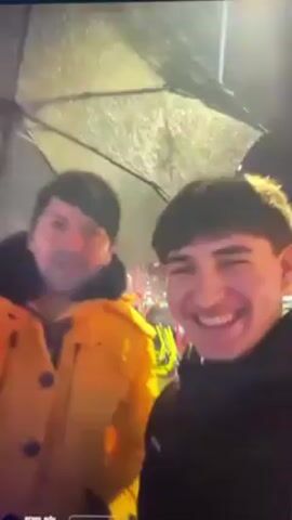 Two men are standing outside in the rain, under an umbrella. The man on the left is wearing a yellow jacket. The man on the right is wearing a black jacket and is holding the camera. Both men are smiling and looking at the camera. There are lights visible in the background, and what appears to be a McDonald's sign is present.
