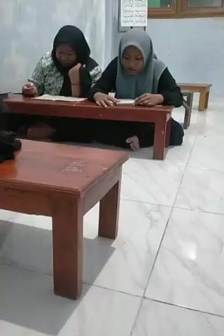 Two young girls wearing hijabs are sitting on the floor in front of wooden desks, reading books. The girl on the left is wearing a black hijab and a patterned top. The girl on the right is wearing a grey hijab and a black top. They are in a room with tiled floors and light-colored walls. There is another wooden desk in the foreground, and a wooden stool or small table in the background. A framed piece of text, possibly religious scripture, hangs on the wall behind the girls.