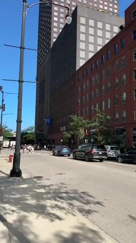 A street view in a city on a sunny day. A tall, brown skyscraper with many windows is in the background, to the left. Next to it is a shorter, brick building with many windows. A black lamppost is in the foreground, to the left. Cars are driving on the street, and there are pedestrians on the sidewalk. The sky is blue and clear.