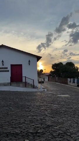 The video shows a car moving down a stone-paved street in a town. The car is dark-colored and approaches the camera. A building with a red door is on the left of the frame with a sign that says "Restaurante Temperodo Rosário". The sky is cloudy with a setting sun, which shines between the building and trees. A baby can be heard cooing.