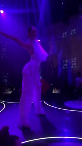 A woman in a white sequined outfit dances on a stage with purple lighting. She is wearing white pants that flare out at the bottom and a white top with a ruffled neckline. Her arms are extended above her head as she performs a dance move. The stage is dimly lit, with purple lights shining down from above. In the background, there are tables with people seated, and some of them appear to be watching the performance. The overall atmosphere is that of a nightclub or a performance venue.