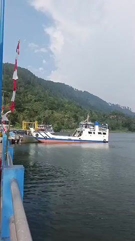 The video opens with a view of a ferry docked at a pier. The ferry is white with blue and orange accents and has a ramp extending from its bow. In the background, a lush green hillside rises from the water, topped with a few scattered buildings. The sky is partly cloudy, with patches of blue visible. As the video progresses, the camera pans to the right, revealing more of the lake and the distant shoreline, which is also covered in dense vegetation. The ferry remains in view for the first few seconds before the focus shifts entirely to the expansive body of water and the horizon. The scene suggests a calm and scenic day on a lake, likely in a tropical or subtropical region, given the verdant landscape.