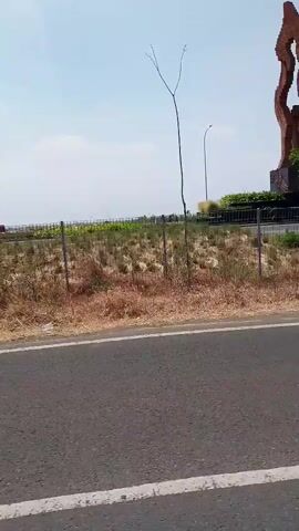 This is a landscape video with a drone perspective of a sculpture that resembles a flame, situated in a dry, grassy area. The sculpture is made of reddish-brown material and stands on a stone base. It is surrounded by sparse vegetation and a metal fence. The background features a clear blue sky with some scattered clouds, a paved road in the foreground, and several tall streetlights and small trees. The camera is moving, suggesting it's either a drone flight or the video is being filmed from a moving vehicle. There are no people or animals visible in the video.
