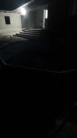 The video captures a dimly lit outdoor scene. In the foreground, the curved edge of what appears to be a large, circular trampoline or swimming pool is visible. The interior of this structure is dark and indistinct. In the background, a building with cinder block walls and a partially constructed roof is illuminated by a bright light overhead, casting long shadows. A set of outdoor stairs leads up to an entrance to the building. The overall atmosphere is dark and somewhat stark.