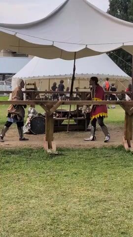 Two individuals, dressed in medieval-style clothing and armor, engage in a sword fight under a large white tent. The person on the left wears a brown tunic and a white head covering, while the person on the right sports a red tunic with yellow trim and similar leg armor. They are positioned on a grassy field, separated by a wooden barrier. Spectators and tents are visible in the background. The fight appears to be a reenactment or demonstration.