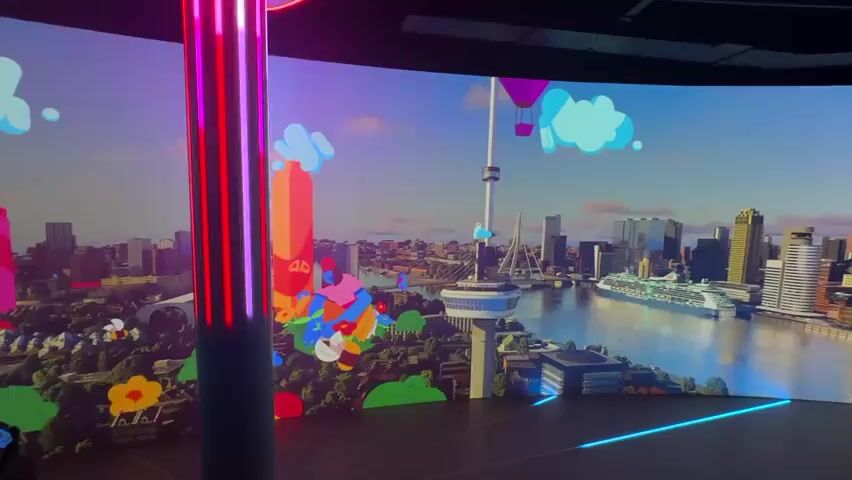 This media file showcases a dynamic, animated digital display of a city skyline, presumably Rotterdam, Netherlands, with a prominent bridge and a tall tower resembling the Euromast. The scene is rendered in a vibrant, cartoonish style with bright colors, exaggerated shapes, and playful elements like hot air balloons and stylized flowers.

The foreground features a vertical column wrapped in pulsing neon lights, shifting from blue to pink and then back again, creating a sense of energy and modernism. Lines of neon lights also radiate from the base of this column onto the dark floor, further enhancing the futuristic ambiance.

Animated characters, depicted in a bold, graphic style, are integrated into the landscape. These figures appear to be interacting with the environment, lounging or playing amongst the stylized trees and buildings. The overall aesthetic suggests a lively, imaginative, and perhaps futuristic interpretation of the city.

The background shows a body of water with a large cruise ship docked. The sky is a gradient of blues and purples, suggesting either a sunrise or sunset, though the stylized nature of the clouds and the overall brightness make it difficult to pinpoint a specific time of day. There are no visible signs of natural weather, but the bright, artificial lighting and vibrant colors evoke a sense of fun and wonder. The emotions conveyed are primarily positive, focusing on playfulness, creativity, and futuristic optimism. No text is visible in the media file.