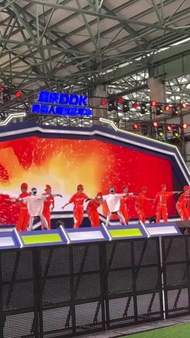 A group of performers in bright orange outfits and white helmets are dancing on a stage in front of a large LED screen. The screen displays dynamic, abstract visuals with colors and effects like fire, smoke, and sparks. The stage is set up indoors with a visible metal roof structure and lighting rigs. A black fence separates the stage from the viewer.