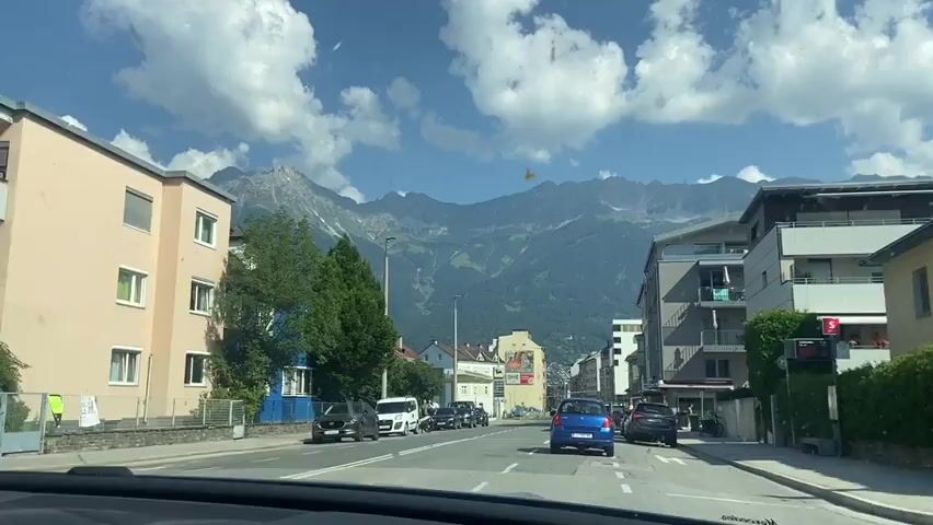 This media file captures a drive through a city in Innsbruck, Austria, under a clear blue sky with fluffy white clouds. The scene is dominated by a wide street lined with apartment buildings and trees. In the distance, a majestic mountain range, possibly the Alps, looms large, its peaks partially shrouded in haze and clouds, indicating a late morning or early afternoon setting.

Several cars are visible on the road, including a blue hatchback, a black SUV, and a white minivan, suggesting typical urban traffic. A gas station with a yellow canopy is prominent on the left side of the street. The architecture of the buildings appears European, with a mix of older and more modern designs. One building on the left is painted a striking blue. A few people are seen walking on the sidewalk, including one individual near a parked scooter.

The overall mood is one of a pleasant, ordinary day in a scenic European city. The bright sunlight and clear skies suggest good weather. There is some visible text on a billboard in the distance, but it is too small to be legible. The view is from inside a vehicle, likely from the driver's perspective, looking forward through the windshield.