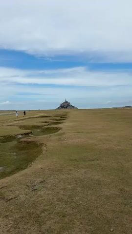A wide-angle video shows a vast, grassy plain under a bright blue sky with scattered white clouds. In the distance, a prominent, medieval-looking castle or abbey sits atop a rocky island. The plain in the foreground features some irregular, shallow depressions in the grass. Two figures, appearing to be people, are walking on the left side of the plain, heading towards the distant structure. One figure is lighter in color and the other darker. The overall impression is one of a scenic, somewhat desolate landscape with a historic landmark in the distance.