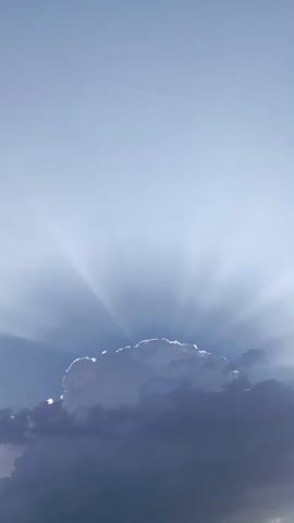 A vertical video shows a cloud formation with sunbeams radiating from behind it. The top half of the screen is a light blue sky, and the bottom half features a large cumulus cloud with darker, denser clouds below it. The sunbeams are visible as shafts of light penetrating through the cloud and into the sky. The cloud is illuminated from behind, creating a bright outline and emphasizing its texture and form. The overall impression is one of a dramatic and ethereal sky scene.