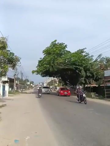 A street view shows several vehicles moving along a paved road. On the left side of the road, there are buildings and utility poles. In the center, a white pickup truck drives past a red car. Further down the road, a large white bus and a dark-colored minivan are visible. A person on a motorcycle is riding in the foreground, facing away from the camera. The sky is partly cloudy, and the scene appears to be set during the daytime.