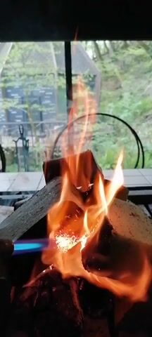 The video shows a close-up of a small fire burning in a fire pit. The fire pit is located on a white tiled surface, possibly a table or countertop, in a covered outdoor area. The camera focuses on the flames as they lick around several pieces of wood arranged in a small pile. A blue flame from a torch is directed at the center of the wood, seemingly being used to start or intensify the fire. Through the glass of the covered area, a blurry view of a green outdoor space with trees and foliage is visible. The lighting in the video is a bit dim, giving the scene a warm and cozy feel. The fire is the central element, drawing the eye with its bright orange and yellow flames.