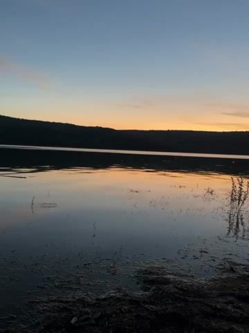 A video clip opens on a body of water with reflections of the sky on its surface. The sky transitions from a deep blue at the top to orange and yellow hues near the horizon. A dark silhouette of hills or a landmass stretches across the horizon line. The foreground shows the murky edge of the water with some debris and vegetation visible. The clip is brief and does not feature any people or significant action. The overall atmosphere is serene and evokes a sunset or sunrise.