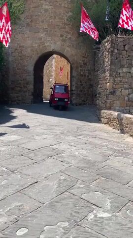 A red three-wheeled vehicle, a Piaggio Ape, drives through an arched stone gateway. The vehicle has a blue object in its bed. Red and white flags hang from poles on either side of the gateway. The ground is paved with large stone slabs. The gateway opens into a street lined with stone buildings and trees. The sky is blue and sunny. The vehicle exits the gateway and turns to the right, disappearing from view.