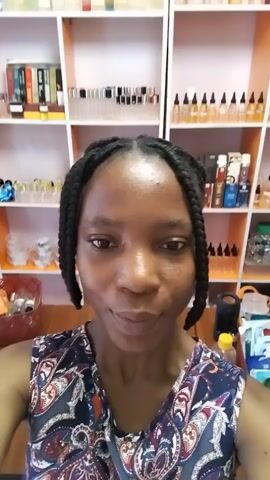 A woman with braided hair is looking at the camera. She is in a room with shelves full of bottles behind her. She smiles at the camera. The video is short and does not contain any dialogue.