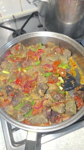 The video shows a close-up shot of food being cooked in a silver-colored metal pan on a stovetop. The pan is filled with various ingredients, primarily chunks of meat and vegetables. The meat pieces are dark brown to greyish-brown and appear to be well-cooked. Mixed in with the meat are vibrant vegetables including thinly sliced red onions, chopped green bell peppers, and small pieces of red bell pepper or tomato.

A black spatula is actively stirring the contents of the pan, mixing the meat and vegetables. There's some liquid or sauce at the bottom of the pan, bubbling gently, indicating that the food is actively cooking over heat. Wisps of steam are visible rising from the pan.

The stovetop has a black grate visible underneath the pan, and part of another pot can be seen in the background to the left. The lighting suggests an indoor setting, likely a kitchen. The overall impression is of a savory dish being prepared.
