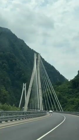 A video captures a white car driving on a highway. The highway is curved and leads towards a large, modern cable-stayed bridge. The bridge has tall, angled concrete towers from which many cables extend to support the bridge deck. The bridge is set in a mountainous region with steep, densely forested slopes on either side. The sky is overcast with clouds. The car is the only vehicle visible on the highway. The camera appears to be mounted inside another vehicle, looking forward and slightly to the right.