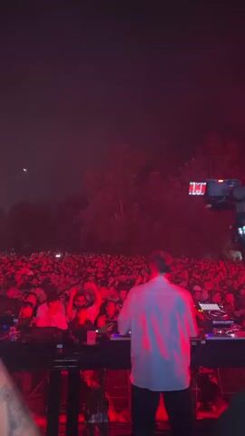 A vertical video opens showing a DJ at a concert. The DJ is wearing a light-colored button-down shirt and dark pants and is standing behind a DJ setup with turntables and mixers. In the background, a large crowd of people is visible, illuminated by red stage lighting that also casts red light on the trees behind them. The crowd appears to be enjoying the performance, with many people raising their hands in the air. The video is dark and grainy due to the low light conditions. The shot is from a slightly elevated perspective, looking down at the DJ and the crowd. A camera on a tripod is visible on the right side of the screen.