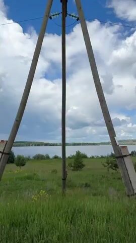 The video shows a scenic view of a body of water, likely a lake or river, with a grassy foreground and a line of trees along the shore. The sky is partially cloudy with patches of blue. Power lines are visible in the upper portion of the frame. The scene appears to be a natural landscape.
