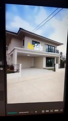 The video displays a digital rendering of a modern two-story house. The exterior shows a light-colored facade, a covered carport, a balcony, and a neatly landscaped yard with a stone pathway. A watermark with the text "Rax Edits" is visible on the front of the house. The interior scenes showcase a living area with a sectional sofa, a coffee table, and a dining area with a table and chairs. The rendering appears to be part of a real estate or architectural visualization project. The video playback controls at the bottom of the screen indicate it is a short clip, possibly a segment from a larger video tour.