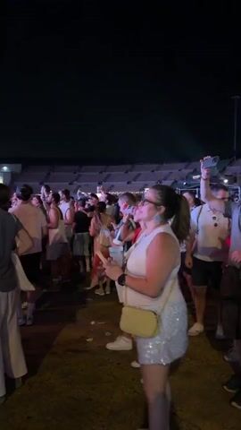 The video begins with a crowd of people at an outdoor event at night. The camera pans to the left, showing a stage with bright lights and a large screen. A banner with the word "vivo" is visible above the stage. The crowd is diverse, with people of various ages and ethnicities. Some people are dancing, while others are watching the performance. The atmosphere appears to be festive and energetic. The video then focuses on a woman with curly hair who is raising her hand in the air. The camera continues to pan across the crowd, showing more people enjoying the event. The lighting is dim, but the stage lights provide a colorful and vibrant backdrop. The overall impression is of a lively music festival or concert.