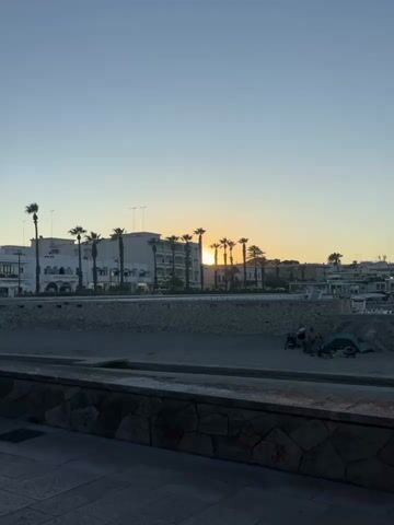 The video shows a beach at sunset. In the foreground, there is a paved walkway with a stone wall. The beach itself is sandy, with some rocks and debris. There is a tent set up on the beach, and two people are sitting near it. Further down the beach, two other people are interacting with the water. In the background, there are buildings with palm trees in front of them. The sun is low in the sky, casting a warm glow over the scene. The water is calm, and the sky is clear.