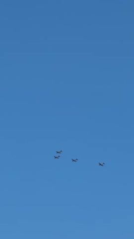 Four small aircraft fly in formation against a clear blue sky. The aircraft appear to be propeller-driven and are spaced relatively close together. The camera is pointed upwards, and the aircraft are visible in the upper portion of the frame. The bottom of the frame shows a hint of a tree-covered hillside, suggesting the footage was taken outdoors on a clear day.