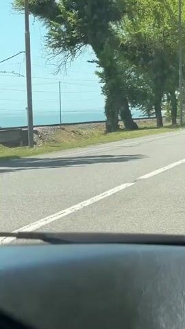 The video captures a daytime view from inside a moving car. The initial shot shows a road bordered by green trees, with a railway track visible behind them, and a body of water (likely the sea) under a clear blue sky in the distance. The perspective then shifts to look forward through the windshield, where a rosary with a cross hangs from the rearview mirror. Vehicles, including a white bus and a grey sedan, are seen driving on the road ahead and approaching from the opposite direction. Trees continue to line the road on both sides. The weather appears sunny. The setting suggests a road trip in a coastal area, likely Primorskoe, Abkhazia, given the context.