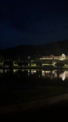 A nighttime scene depicts a large, illuminated building on a hillside across a dark body of water. The building is long and appears to be a resort or hotel, with many windows and decorative lights outlining its structure and the surrounding pathways. The lights from the building reflect on the water's surface. Trees are visible between the water and the building. The sky is dark, indicating night. The foreground shows a dark grassy area and a concrete barrier.