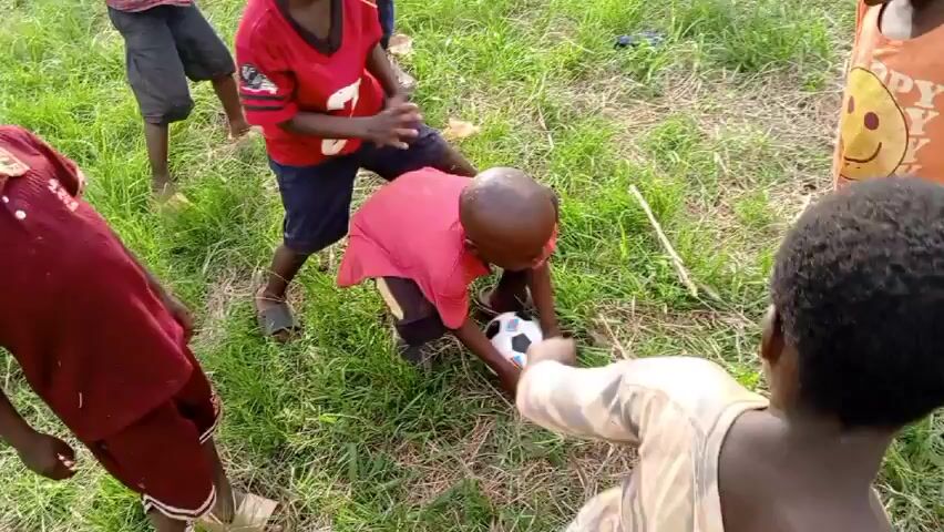 A video depicts a group of approximately a dozen young children, likely in an African or similar rural setting, interacting on a grassy field. The children are of varying sizes and complexions, primarily wearing casual or worn clothing and sandals. Several are barefoot. One child wears a patterned shawl-like garment. A soccer ball is visible, held and passed between some of the children. Some children are shouting and jostling for the ball. The camera angle is from a low vantage point, likely held by one of the children or an adult close to the group. The overall environment is a grassy area with some taller grass in the background.
