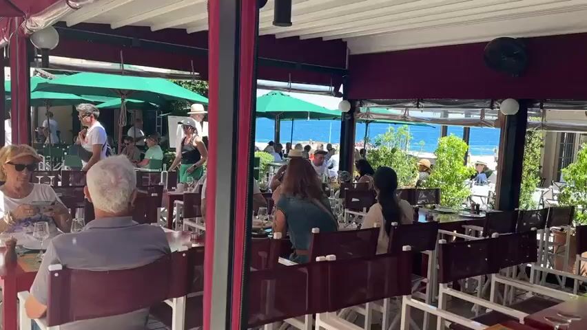The video shows an outdoor dining area of a restaurant with people seated at tables. The tables are covered with red tablecloths and have white chairs. In the background, there is a man playing a guitar and another man standing. There are also green umbrellas providing shade. Beyond the restaurant, the sea is visible. Some of the people in the foreground are looking at their phones. The overall atmosphere appears relaxed and social.